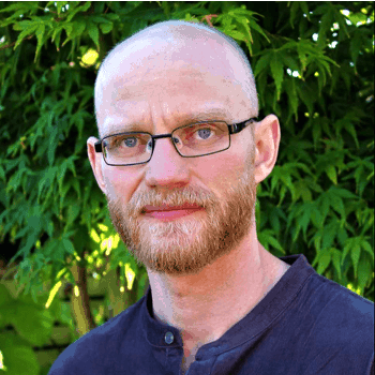A man in a dark blue t-shirt stands in front of a tree