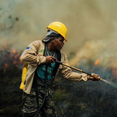 A man in a yellow hard hat uses a hose to extinguish a fire in a grassy area