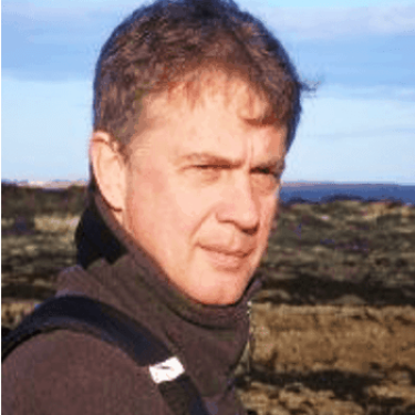 A man in a grey fleece stands in front of a rural landscape