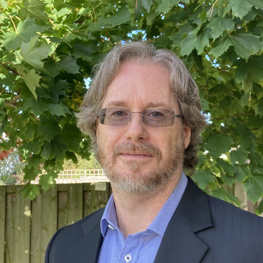 A man in a blue shirt and dark blue jacket stands in front of a tree