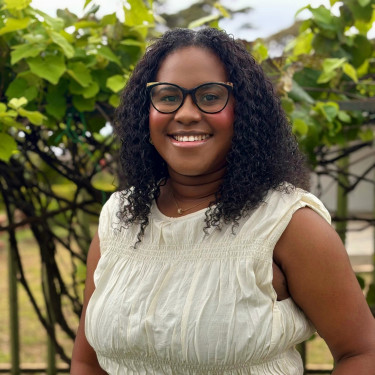 A woman in a white top stands smiling in front of a tree