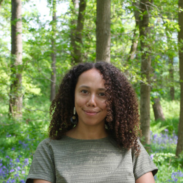 A woman in a grey t-shirt stands smiling in a wood with bluebells behind her
