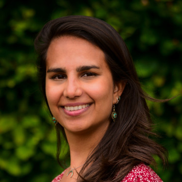 A woman in a red floral top stands smiling in front of leaves