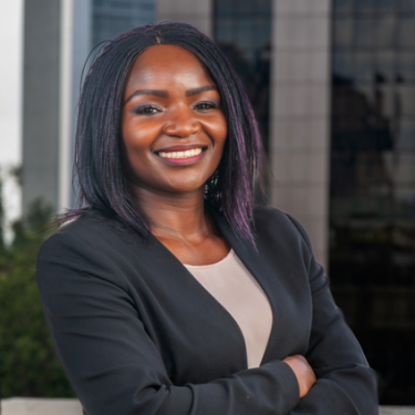 A woman in a white top and black jacket stands smiling in front of a window