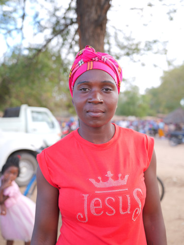 A woman in a pink headscarf looks at the camera with a tree and street behind her