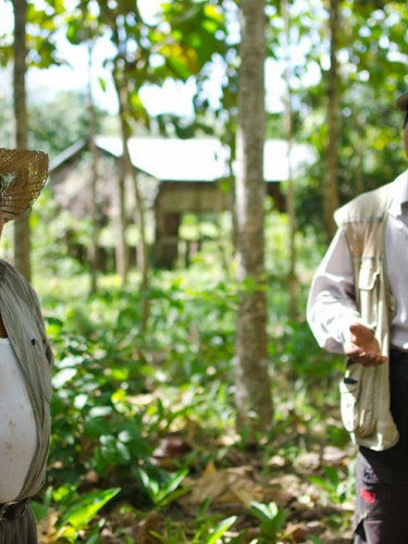 Two men in hats stand talking in a wood with a house in the background