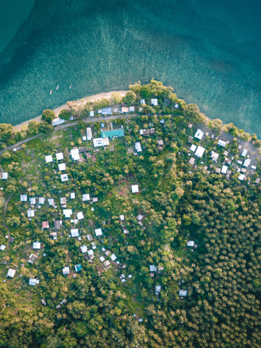 Aerial view of a coastal village with scattered houses, surrounded by dense greenery, and bordered by clear blue water.