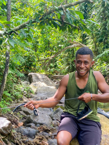 A man in a green vest holds spear-fishing gear by a river in a forest