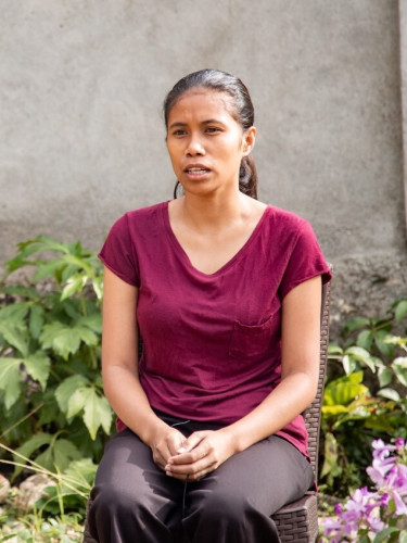 A woman in a fuchsia t-shirt sits in front of a wall and some plants