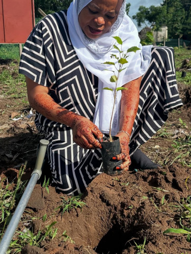 A woman in a hijab kneels to plant a seedling with a crutch on the ground next to her