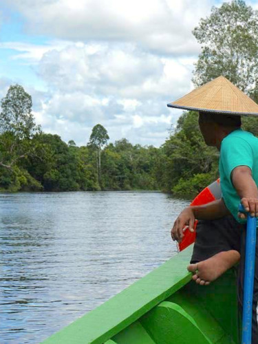 A man in a traditional straw hat sits facing away from the camera on the edge of a boat navigating a river with forested banks