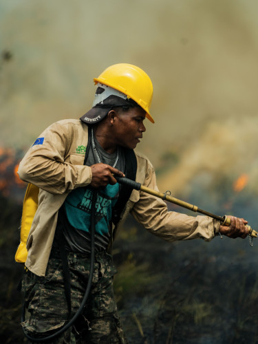 A man in a yellow hard hat uses a hose to extinguish a fire in a grassy area