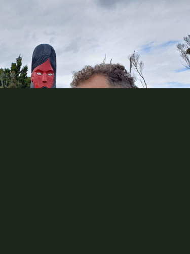 A man in a grey t-shirt stands next to a red carving of an ancestral figure
