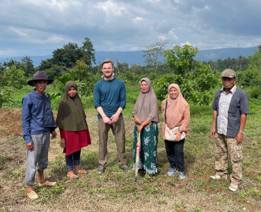 A group of six people stand alongside each other in the sun in a rural tropical setting