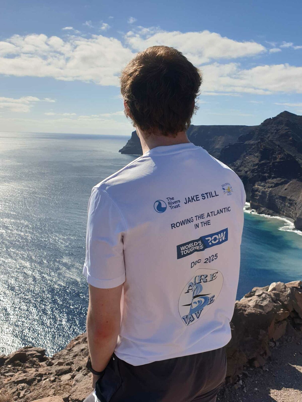 Photo from behind of a man stood overlooking a bay of water from a cliff.