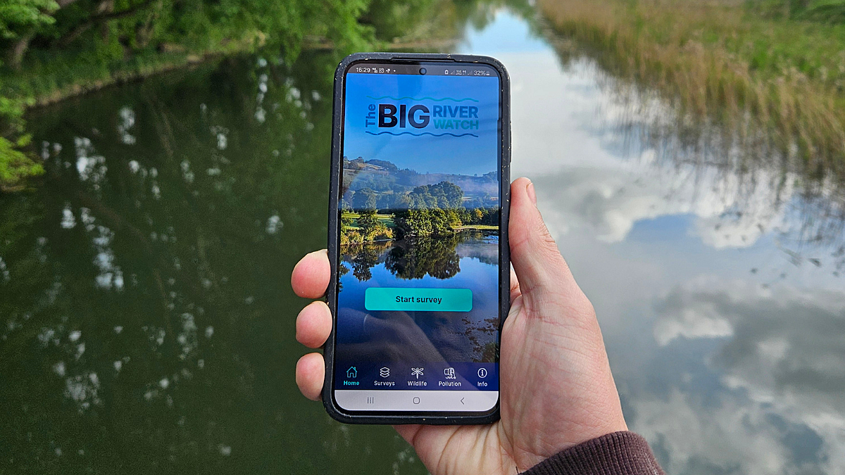 Hand holding a phone in front of a river, displaying the Big River Watch app