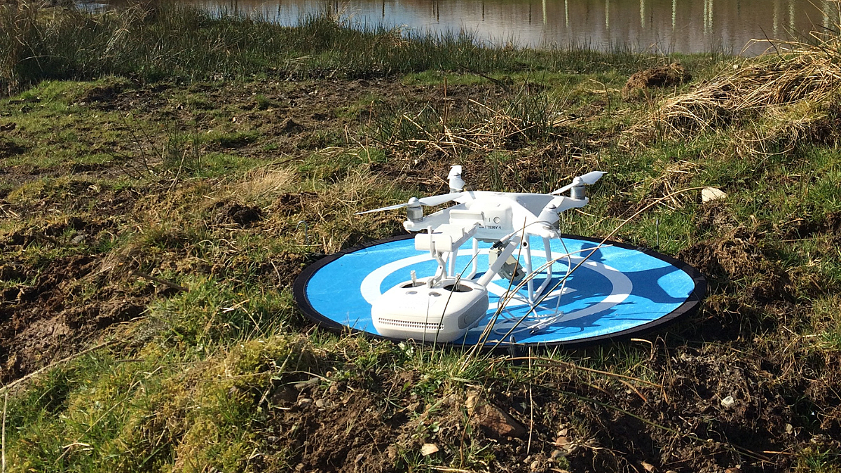 White drone sat on blue landing pad in field with water in background