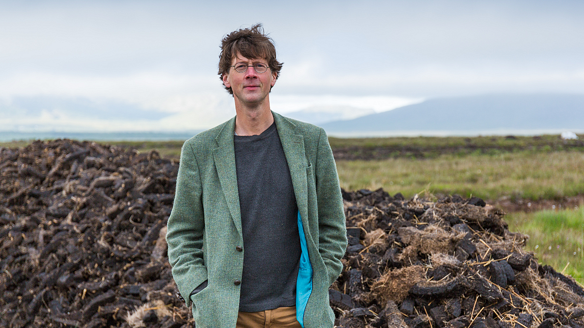 White man stood outside with grass behind him and blue sky