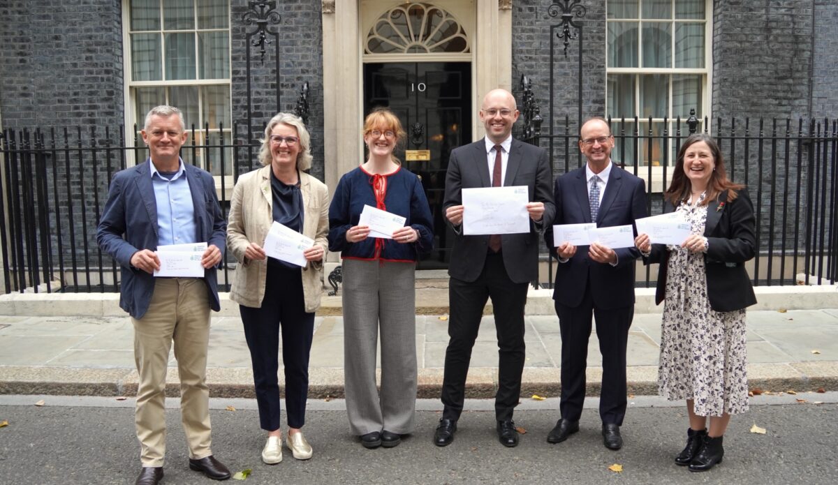 Group of six people, three women and three men stood outside a building with a black door holding envelopes