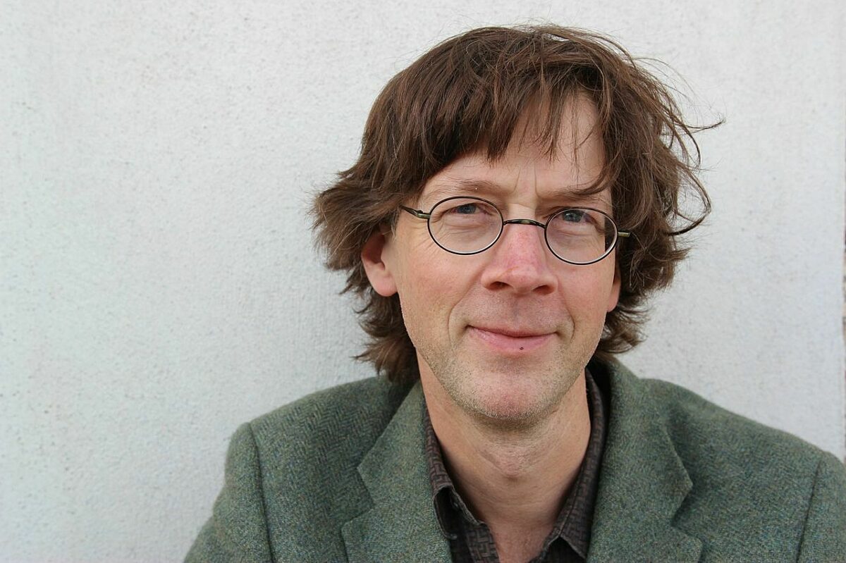 A man looks at the camera, with round glasses and brown hair