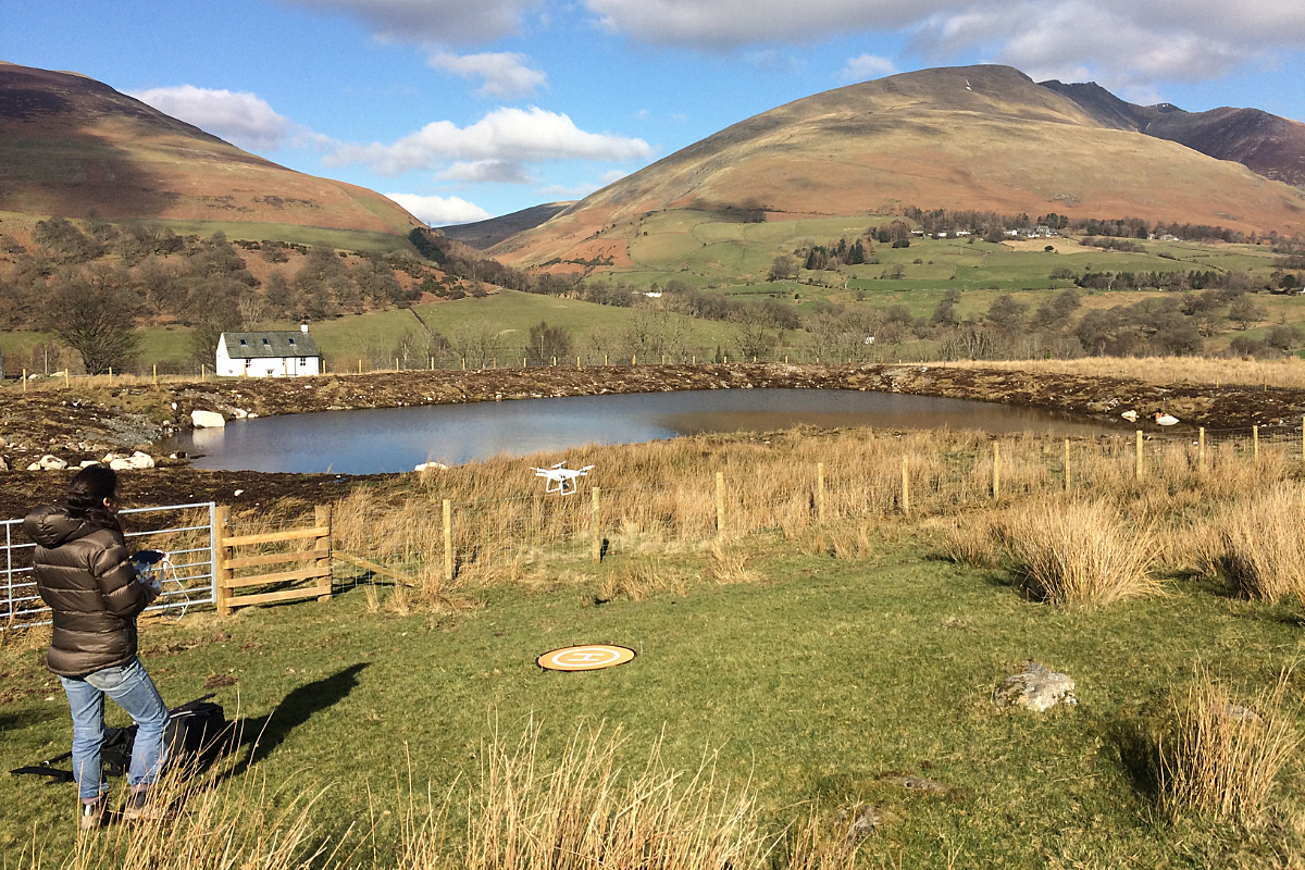Woman in jeans and black coat stood in field looking at a lake with mountains in background flying a white drone
