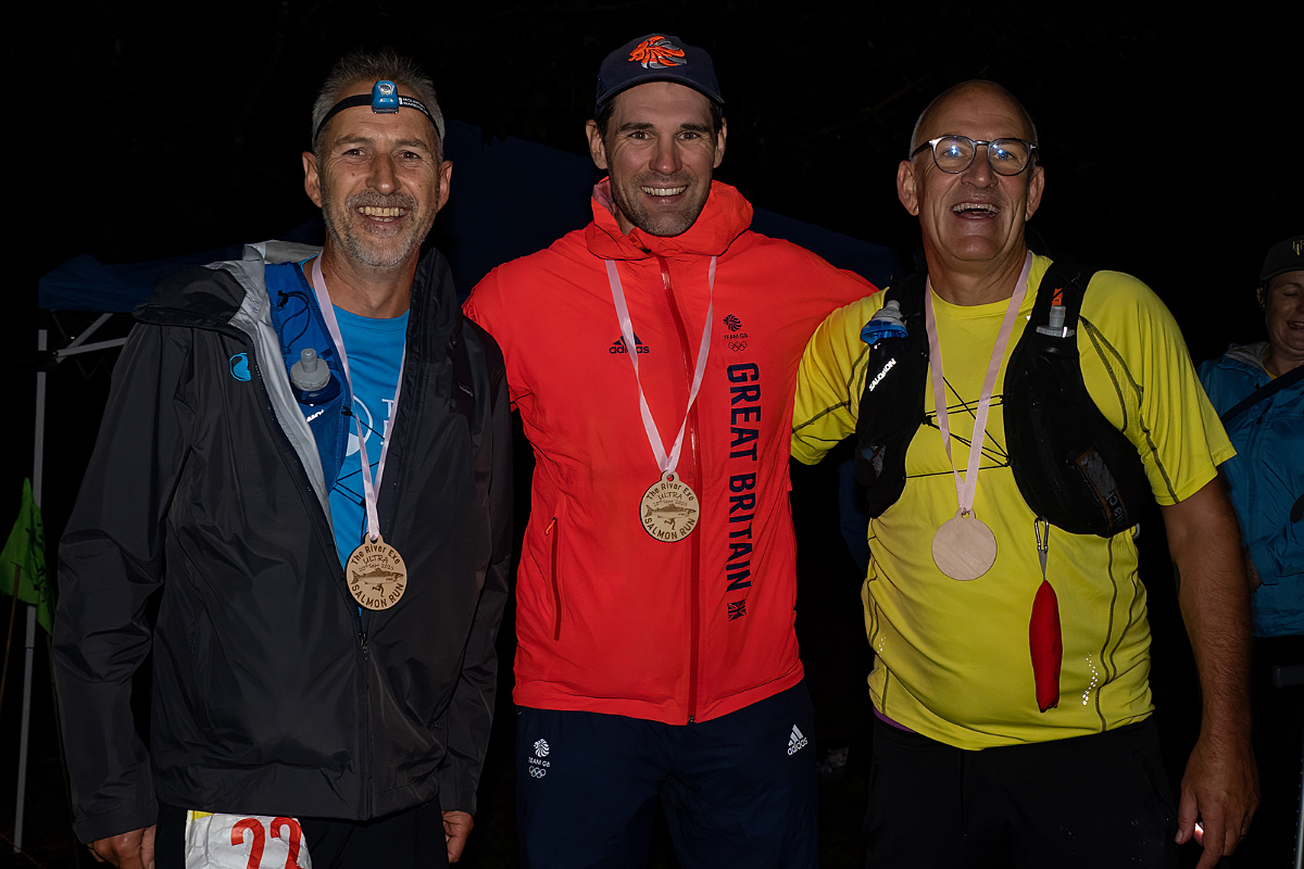 Three male runners stood arm in arm in the dark with medals around their neck
