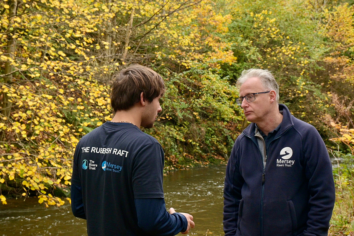 Young male with brown hair in navy blue t-shirt on the bank of a river talking to an older male in a dark blue jumper.