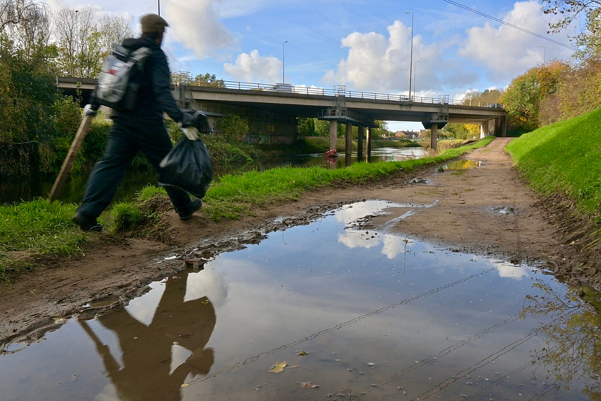 Young male in black coat, wearing a rucksack, walking alongside a river holding a rubbish bag and a litter picker