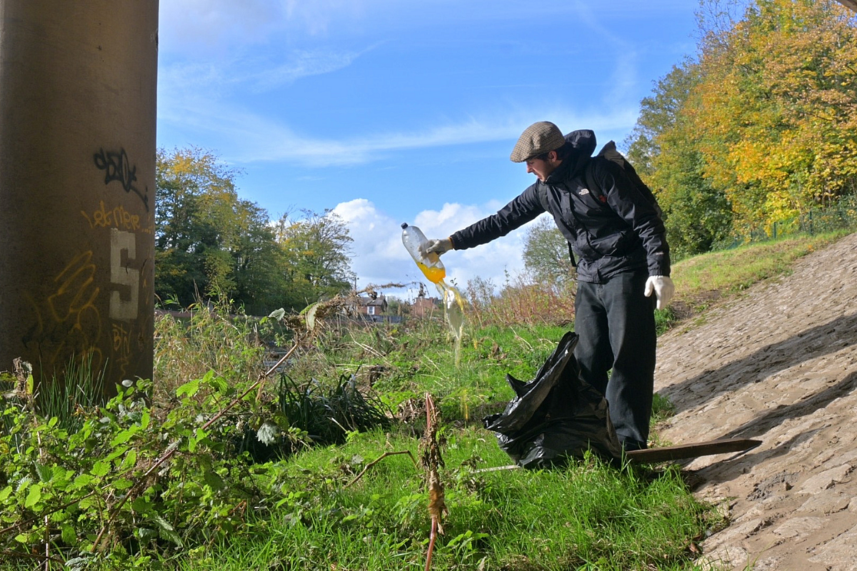 Young male in black coat and flat cap on a grassy bank holding up a plastic bottle filled with dirty water