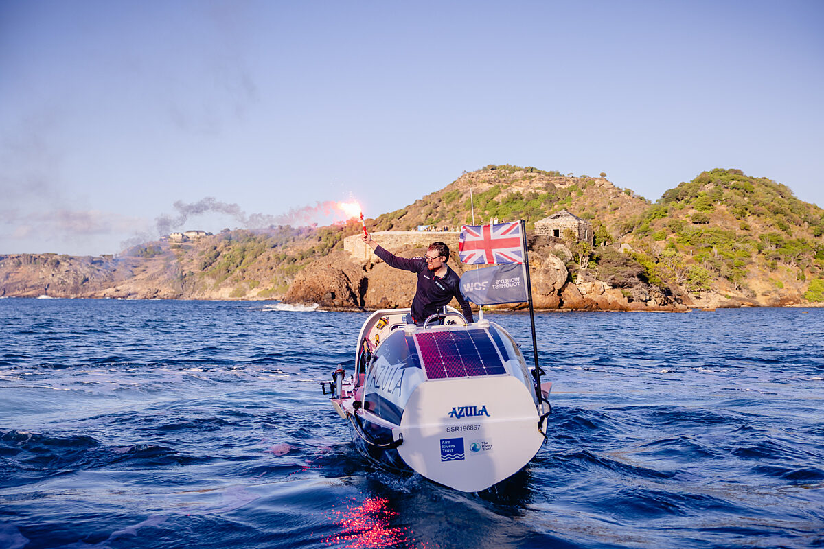 Man on white rowing boat holding a red flare on the water with land behind him