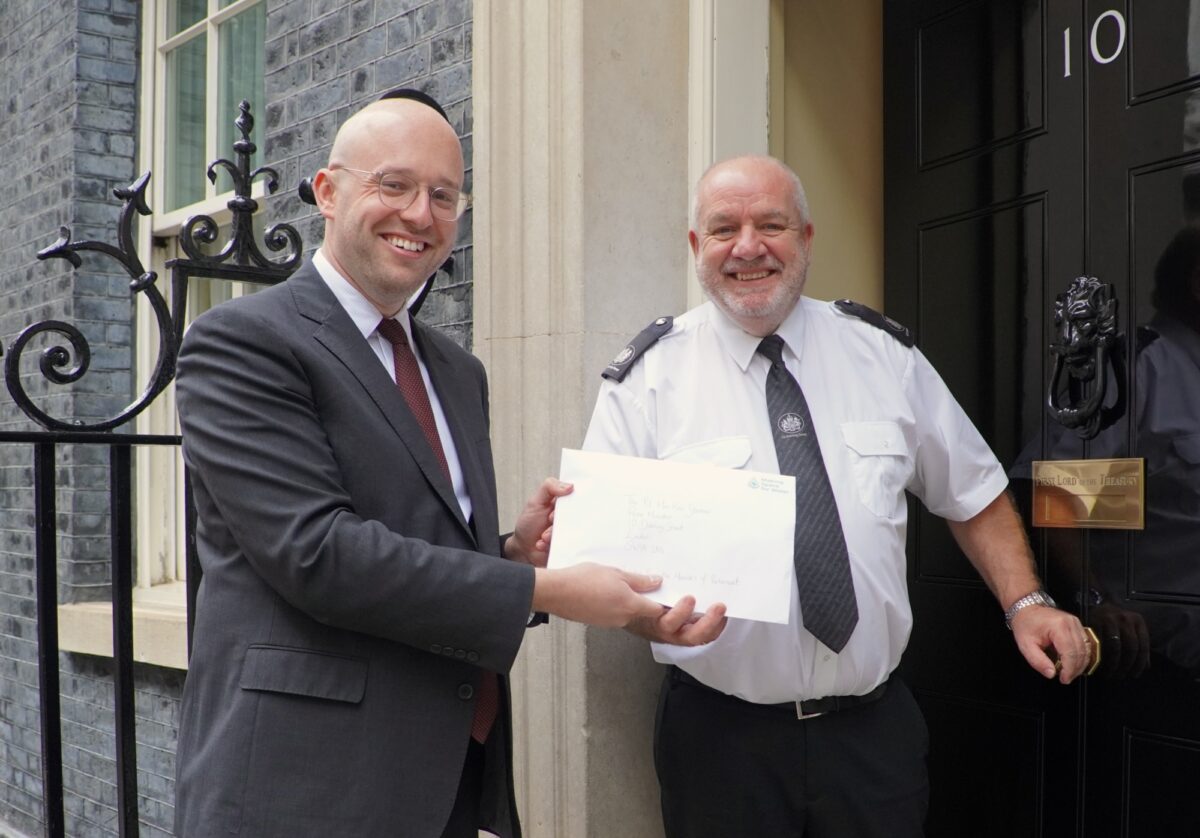 Bals white man in black suit and red tie handing a letter to a bald white man in white shirt and black tie in front of black door with number 10 on it