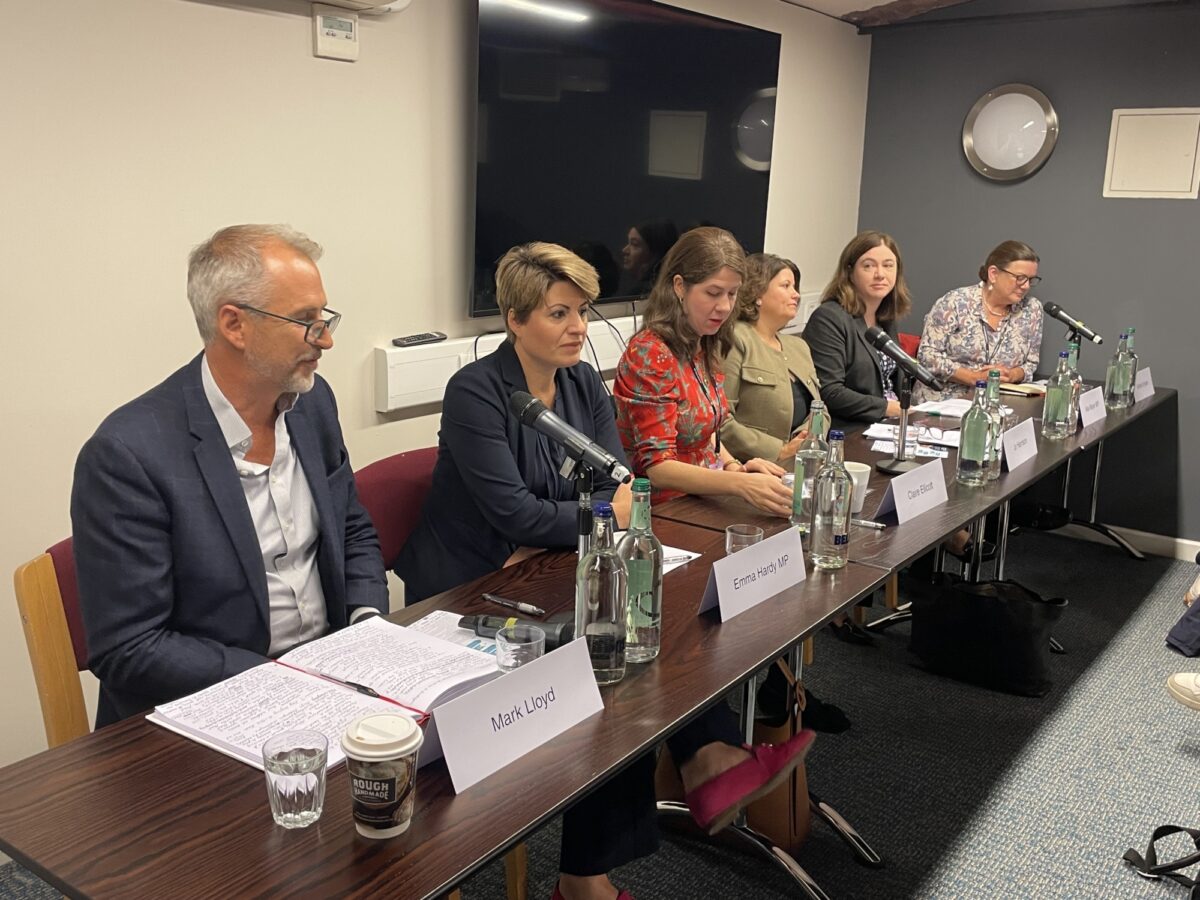 Five speakers on a panel sit at a long desk facing a room