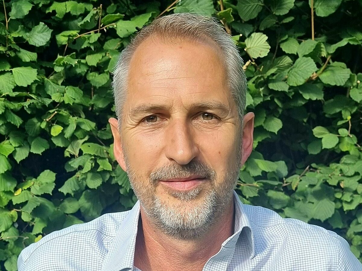 Image shows a man looking at the camera against a backdrop of green leaves