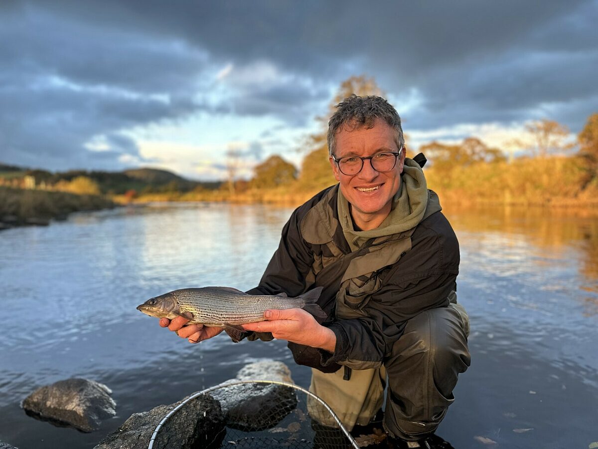 A smiling man holds up a fish by the bank of a river in sunlight
