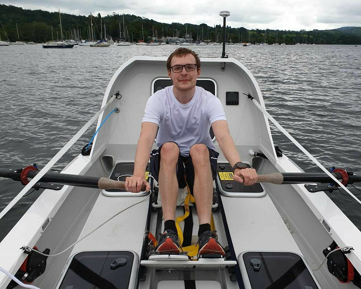 A man in a white teeshirt looks at the camera as he rows a large white boat solo on a lake