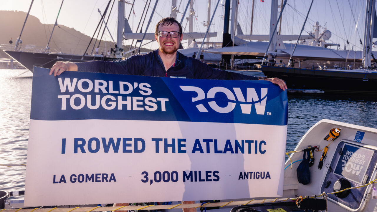 Man smiling holding banner that says I rowed the Atlantic, La Gomera, 3000 miles, Antigua