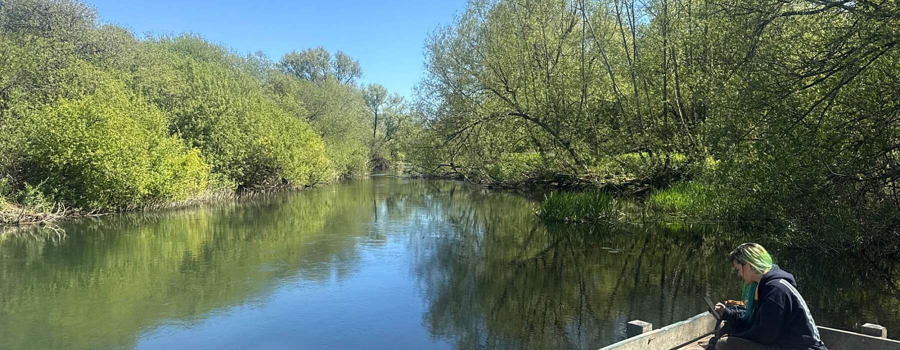 Woman with green hair sat crossed legged on a wooden jetty over a river.