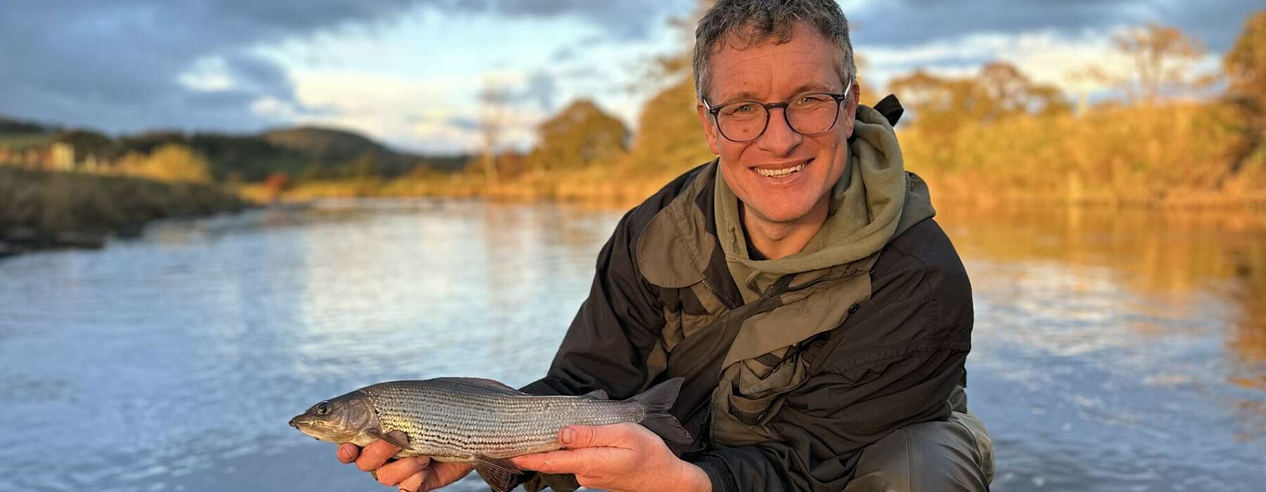 A smiling man holds up a fish by the bank of a river in sunlight