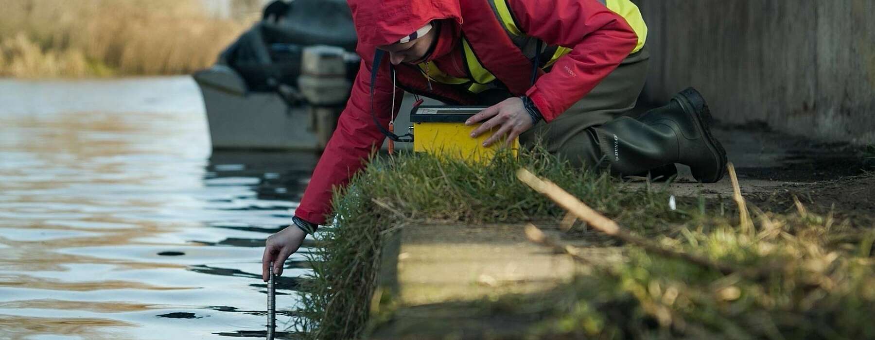 Person in red hooded raincoat and yellow high visibility jacket leaning over a river dipping a test tube into the water