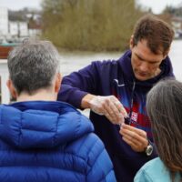 A man with brown hair in a blue hoodie wears plastic gloves while testing a sample of river water for nitrates