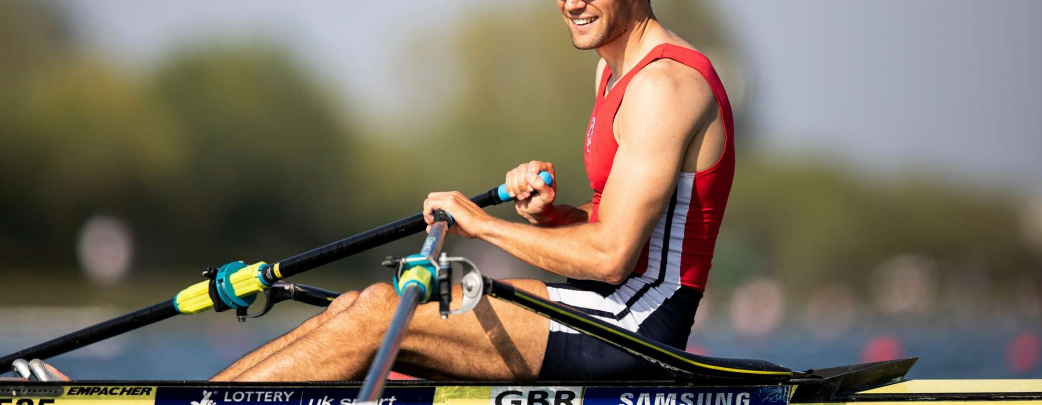 A smiling male rower sits in the single rowing boat, holding his oars in the sunshine