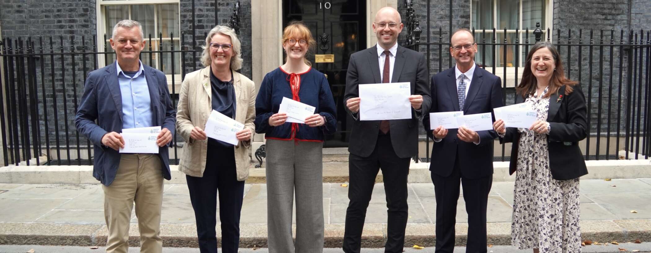 Group of six people, three women and three men stood outside a building with a black door holding envelopes