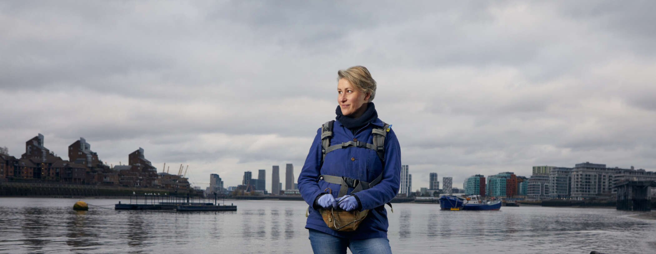 A smiling woman stands on the banks of the Thames, looking into the distance