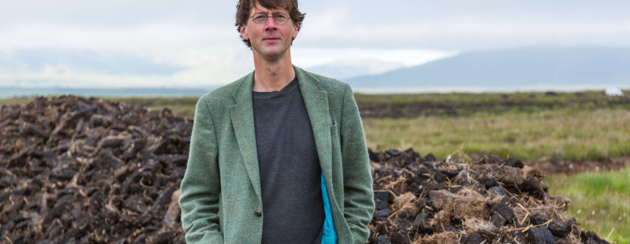 A smiling man looks at the camera, with round glasses and brown hair. In the background is countryside