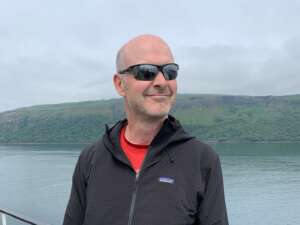 A smiling man in sunglasses stands in front of a lake