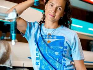 Rower Imogen Grant stands in front of rowing boats, smiling and wearing a light blue teeshirt with a dark blue bold river print