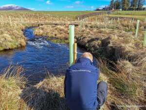 Man in blue waterproof crouched by a river planting trees