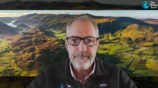 White man with beard and black glasses sat in front of an image of a landscape