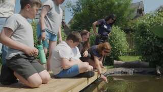 Children kneeling next to a pond with an adult woman pointing to something in the water
