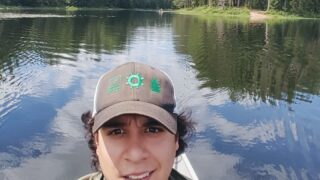 A smiling man in a hat sits on a boat in a calm canadian river